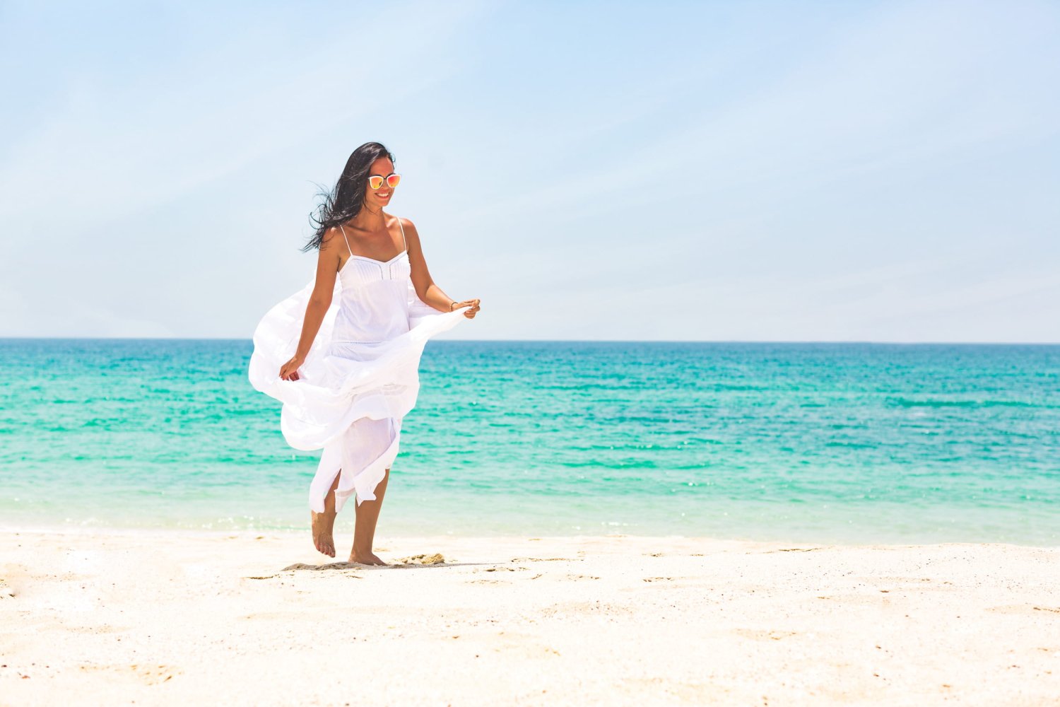 A smiling woman in a flowing white dress and sunglasses walks barefoot along a pristine white sand beach. The bright turquoise ocean and clear sunny sky behind her perfectly capture the carefree, relaxing 