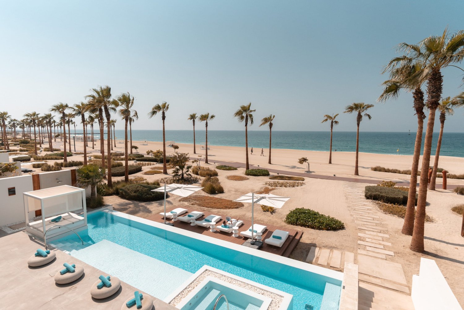 An elevated view of a luxurious private pool area at Nikki Beach Resort & Spa Dubai, featuring a bright blue swimming pool with submerged loungers, a white cabana bed, and a wooden deck with white sunbeds and umbrellas. Tall palm trees line the sandy path leading directly to the wide beach and ocean under a clear sunny sky, capturing the ultimate feel-good staycation escape on Pearl Jumeira