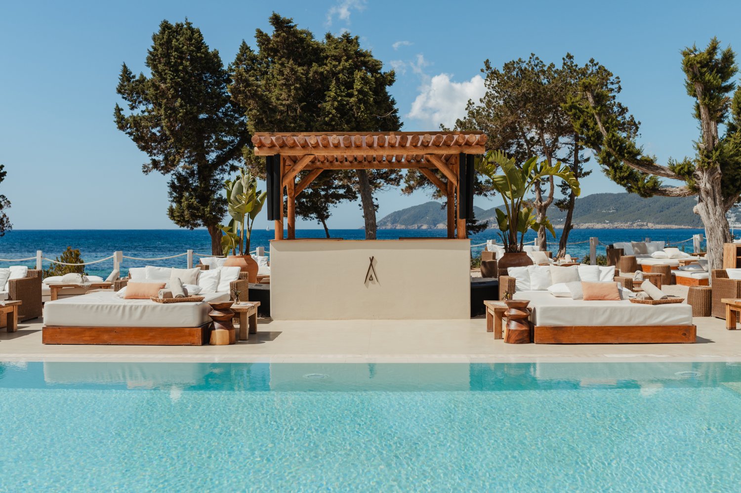 A serene view across a pristine swimming pool at Nikki Beach Ibiza, centering on a wooden pergola DJ booth adorned with the brand's signature teepee logo. Flanked by luxurious white wooden daybeds overlooking the vibrant blue sea and distant coastline of Santa Eulalia, this sun-drenched scene perfectly captures the refreshed look and daytime beats of the Balearic favorite