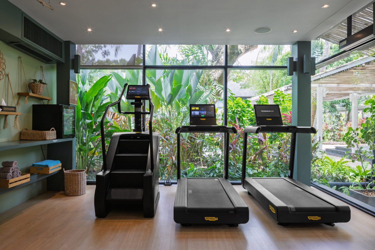 A bright fitness center featuring two modern treadmills and a stair climber machine. The equipment faces a wall of floor-to-ceiling glass windows that provide a calming, immersive view of a dense tropical garden