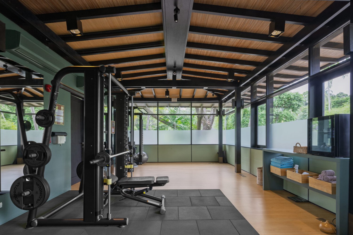 A modern, serene resort gym featuring a multi-functional Smith machine for strength training. The space includes light wood floors, calming green walls, a dark beamed ceiling, and a hydration station with fresh towels, all illuminated by natural light from large windows looking out into lush tropical greenery