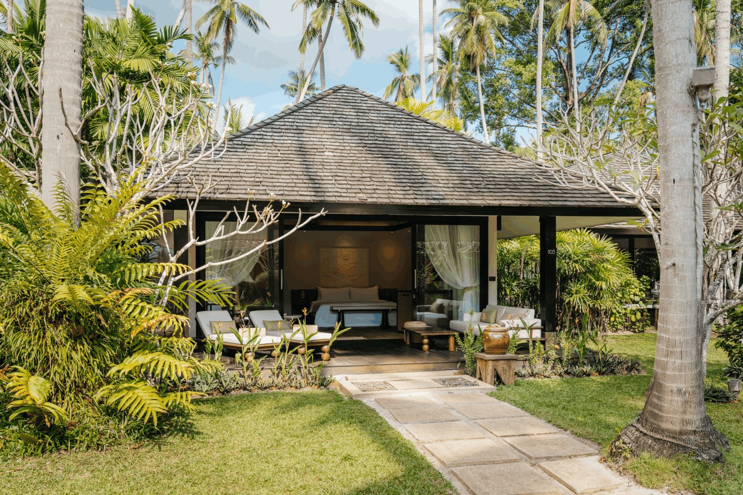 Wide exterior view of the new Signature Palm Villa at Nikki Beach Koh Samui, nestled among towering coconut palms and lush tropical ferns. A stone pathway leads to a private wooden deck furnished with modern white lounge chairs, while large open glass doors reveal the luxurious bedroom interior, blending traditional Thai roof architecture with contemporary design