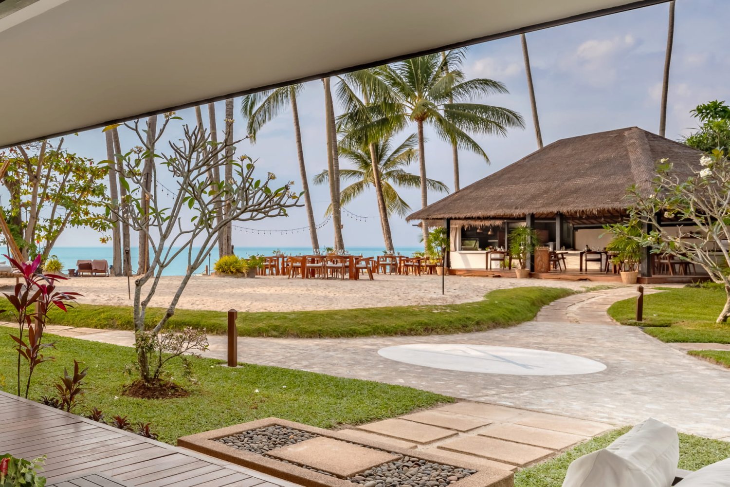 View from the terrace of a Signature Palm Villa looking out towards the sandy Lipa Noi Beach, featuring outdoor dining tables set under towering palm trees and a thatched-roof beach pavilion, with the blue ocean visible in the distance