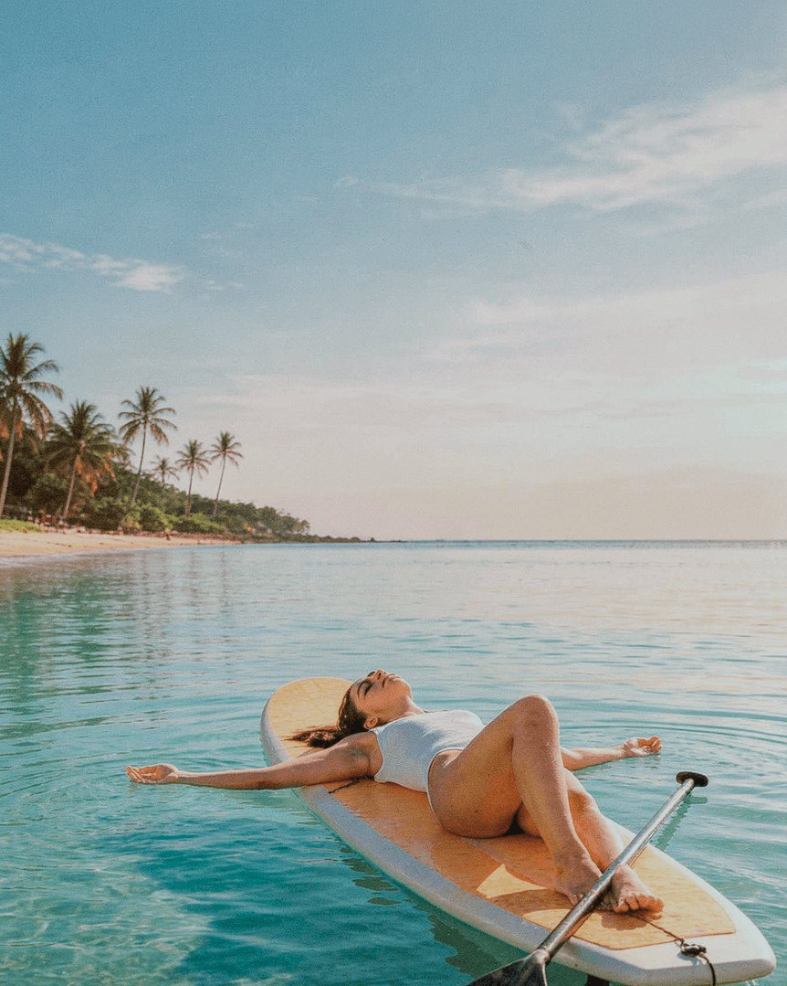 A woman relaxing in a shavasana pose on a paddleboard, floating on calm turquoise waters with a palm-lined beach in the background, capturing the essence of a peaceful wellness session