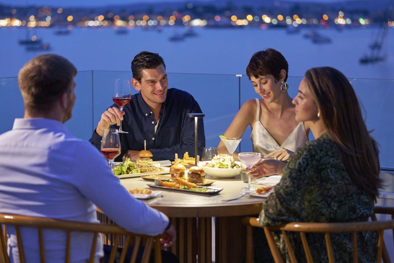 A group of four friends enjoying an elegant evening meal together at a round table on a glass-balcony terrace, overlooking a serene bay with twinkling city lights in the background. They are smiling and sharing wine, cocktails, and a variety of dishes including salads and casual bites, perfectly capturing the vibrant Mediterranean dining experience at Porto Heli