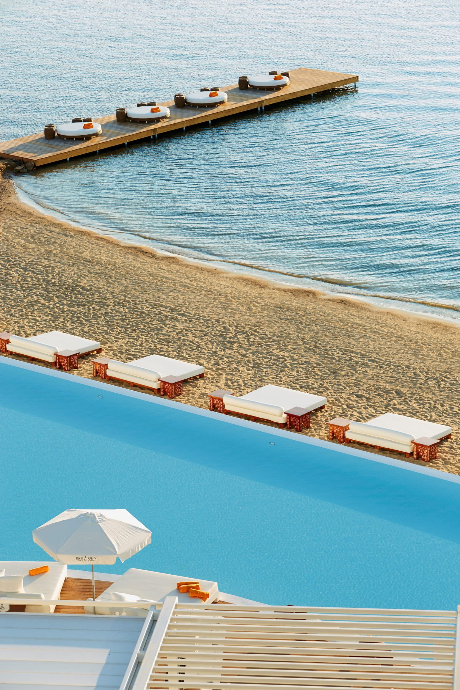 High-angle view of the infinity pool and sandy beach at Nikki Beach Resort & Spa Porto Heli, featuring a wooden pier extending into the calm blue sea lined with luxury sunbeds