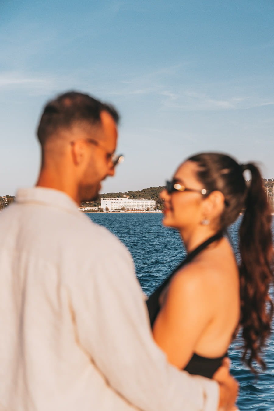 A blurred view of a couple in the foreground embracing by the water, framing a sharp, distant view of the white Nikki Beach Resort & Spa Porto Heli building across the sparkling blue sea. This romantic, sunlit scene beautifully captures the allure of extending your getaway to enjoy up to 20% off on stays of five nights or more