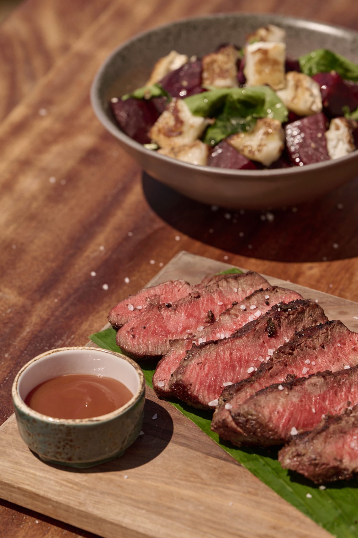 A close-up view of perfectly sliced medium-rare steak seasoned with flaky sea salt, beautifully presented over a green leaf on a wooden board alongside a small bowl of savory dipping sauce. With a fresh salad of roasted beetroot and grilled cheese blurred in the background on a rustic wooden table, this appetizing scene highlights the premium, fresh local ingredients featured on the Nikki Beach Porto Heli dinner menu