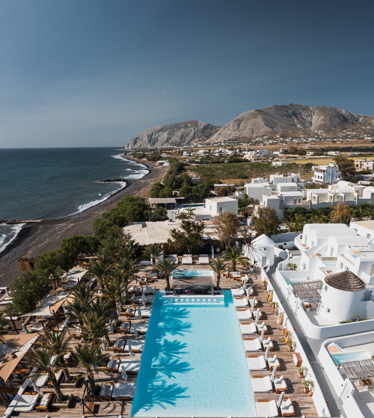 A breathtaking aerial shot of Nikki Beach Santorini, showcasing the expansive swimming pool lined with luxurious white daybeds, the distinctive Monolithos black sand beach, and the dramatic coastal mountains along the edge of the Aegean Sea
