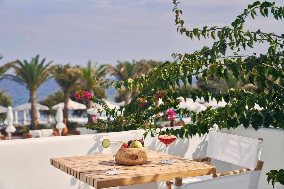 A sun-drenched balcony framed by vibrant bougainvillea branches, featuring a wooden table set with two crafted cocktails and a bowl of fresh fruit, with palm trees and the ocean in the background