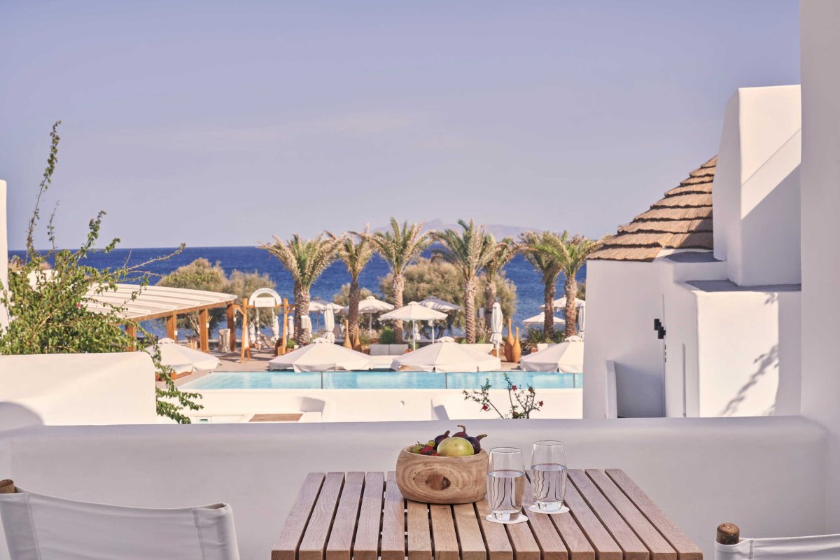A serene balcony view featuring a slatted wooden table set with fresh fruit and water, looking out over a luxurious swimming pool lined with palm trees and the sparkling Aegean Sea
