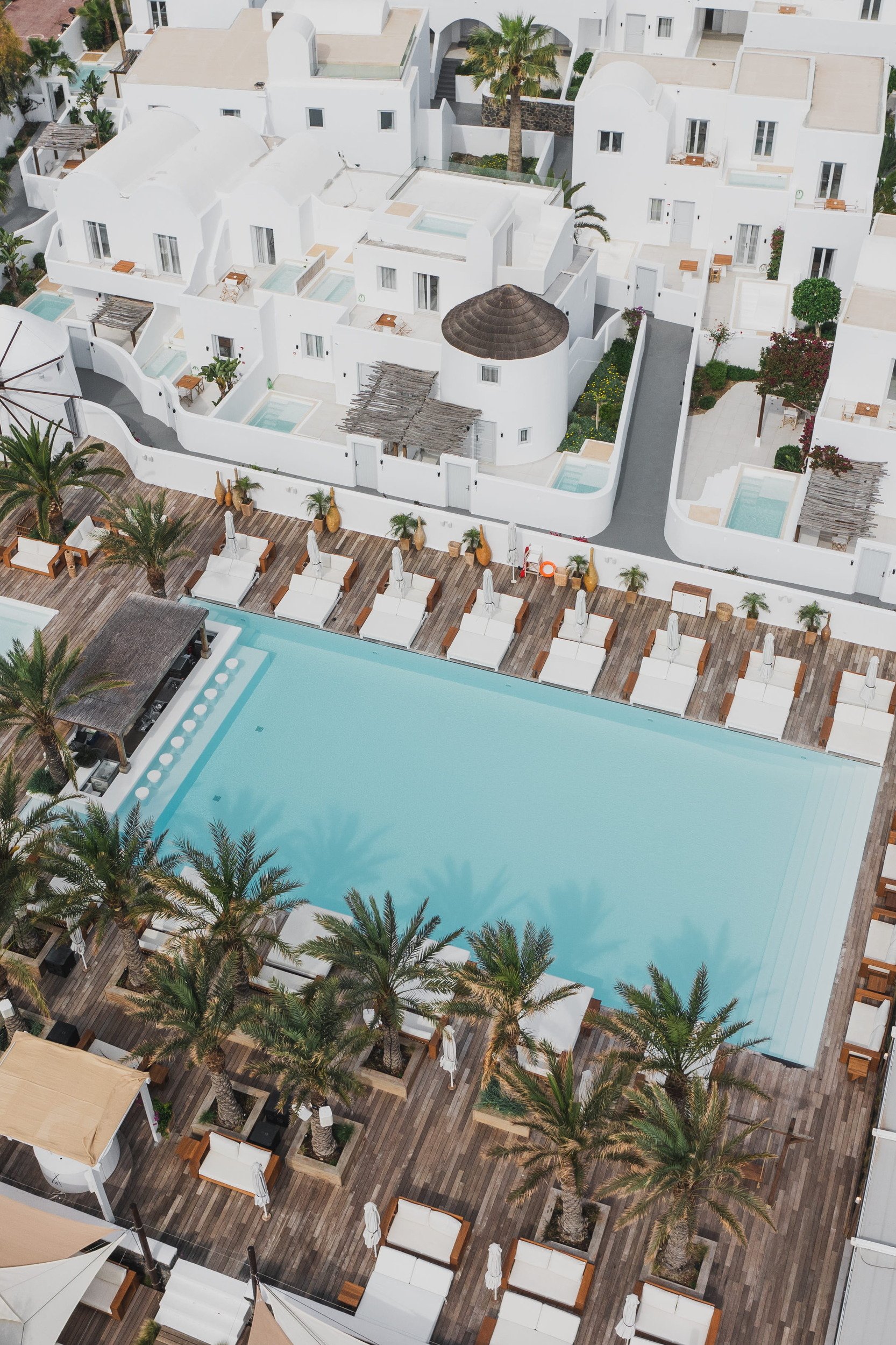 An aerial view of the Beach Club at Nikki Beach Resort & Spa Santorini, showcasing the brand's signature barefoot luxury ambiance. A large, pristine swimming pool featuring a swim-up bar is surrounded by an expansive wooden deck lined with plush white daybeds and towering palm trees. In the background, traditional all-white Cycladic architecture with private plunge pools perfectly captures the island's stunning aesthetic