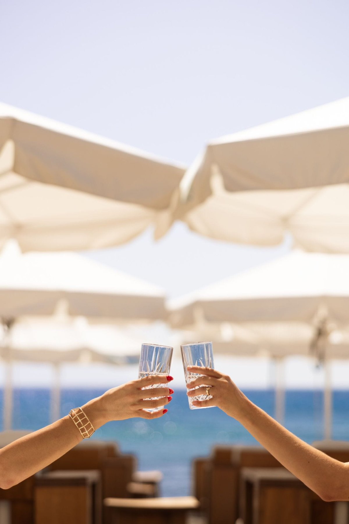 Two hands clinking glasses of ice water in a refreshing toast beneath white parasols, with the sparkling blue Aegean Sea in the background. This beautifully captures the relaxing, unforgettable atmosphere of dining and unwinding at Nikki Beach Santorini