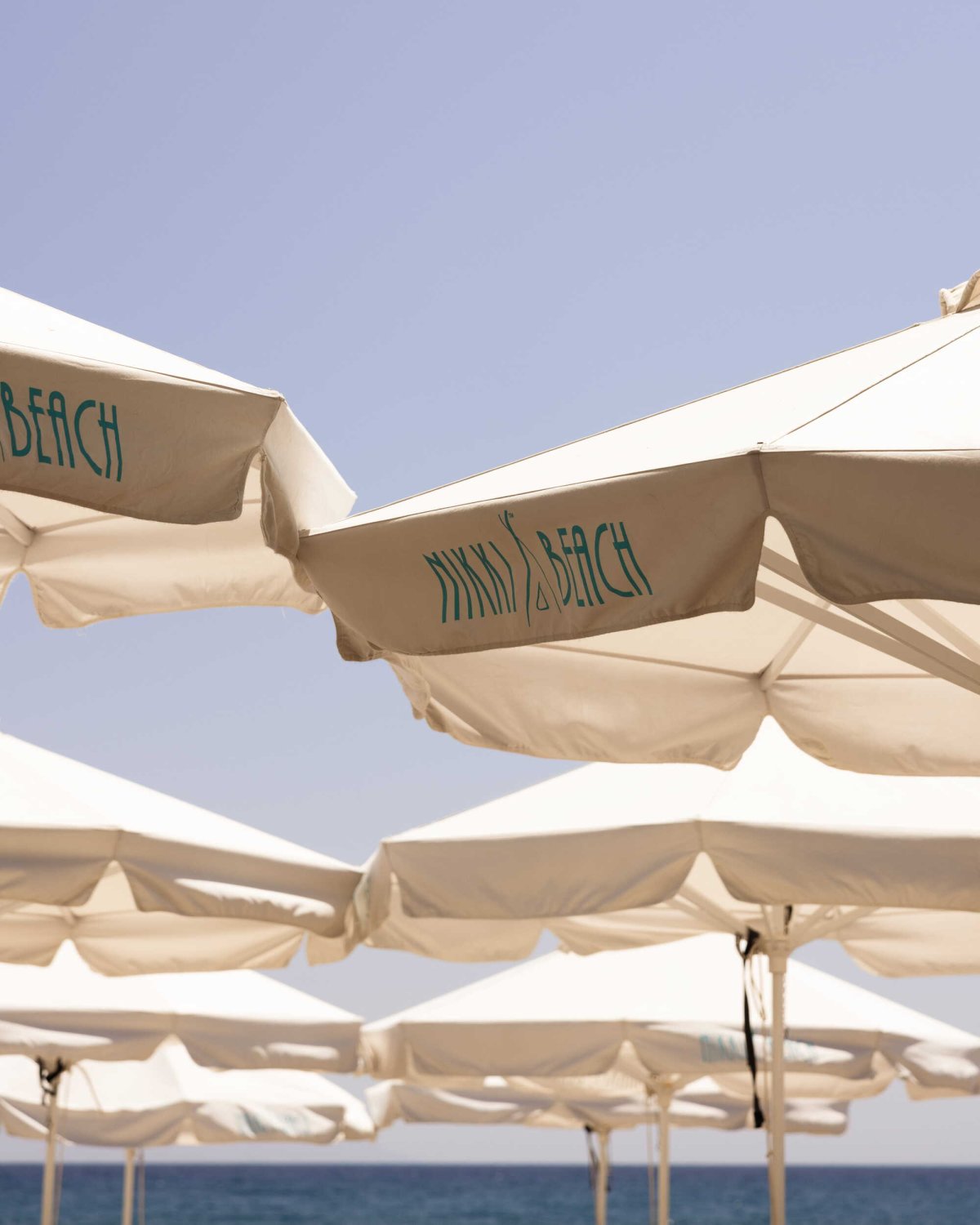 A cluster of classic white parasols featuring the teal Nikki Beach logo, set against a clear blue sky and the deep blue waters of the Aegean Sea