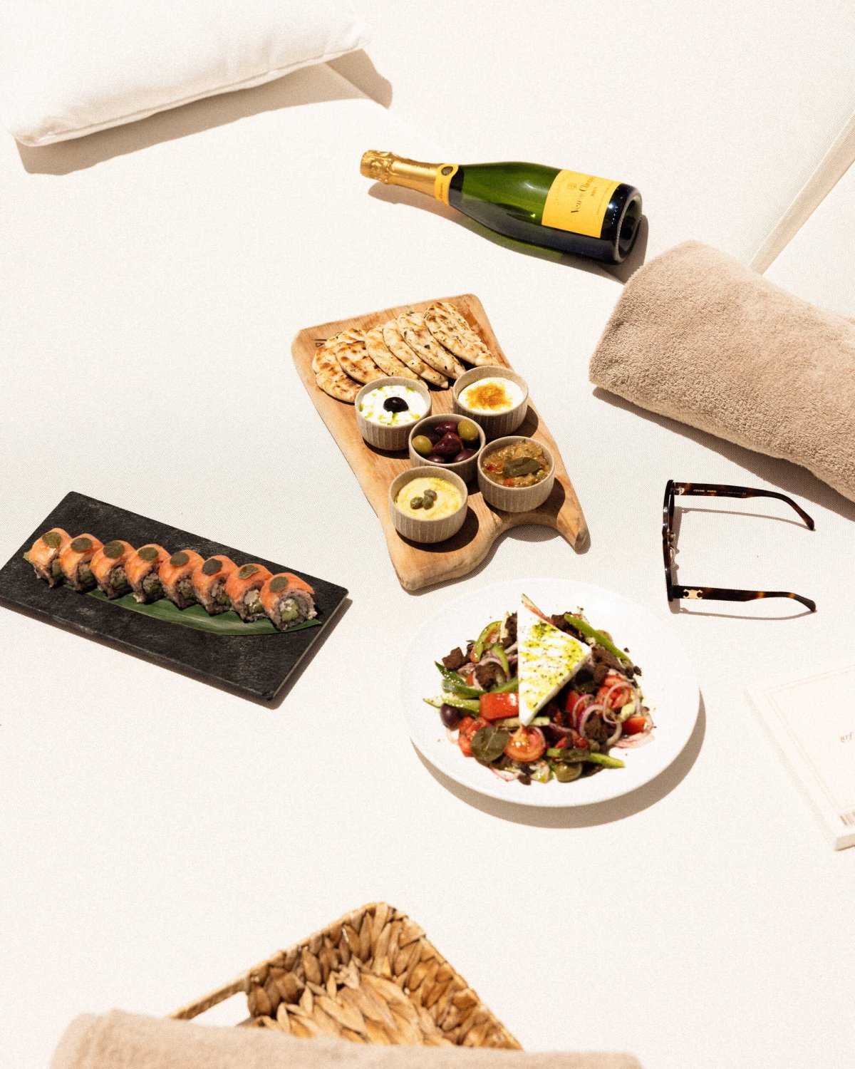A top-down view of an exquisite sharing menu laid out on a plush white daybed. The spread includes a fresh Greek salad, a wooden mezze board with pita and dips, sushi rolls, and a chilled bottle of champagne, perfectly embodying the globally inspired coastal cuisine