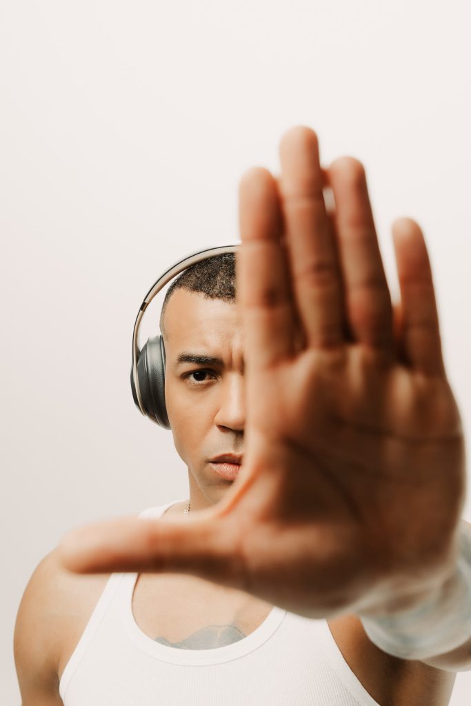 A striking studio portrait of special guest DJ Steve Provis wearing over-ear headphones and a white tank top. He looks intensely at the camera while holding his hand out toward the lens, partially obscuring his face. This bold image perfectly captures the dynamic energy he will bring to his set at the "Celebrate The Sun" season opening event at Nikki Beach Santorini