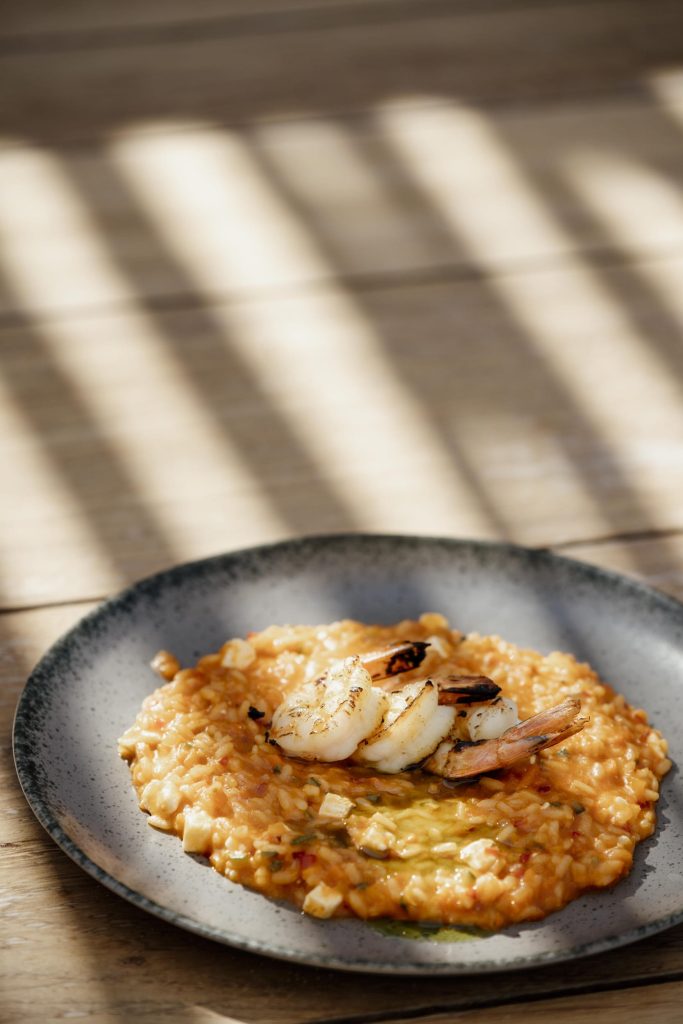 A close-up view of a vibrant Mediterranean seafood dish featuring a rich, tomato-based risotto topped with perfectly grilled shrimp, cubes of artisanal Greek feta cheese, and a drizzle of fresh olive oil. Served on a dark ceramic plate resting on a sunlit wooden table with dramatic pergola shadows, this mouth-watering plate beautifully represents the authentic Greek gastronomy and fresh Aegean seafood curated for the new Amazing Sundays - Greek Tables dining series at Nikki Beach Santorini