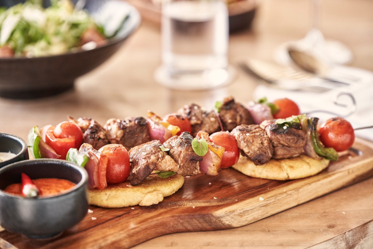 A close-up view of appetizing grilled meat and vegetable skewers resting on warm flatbreads, presented beautifully on a rustic wooden board alongside small bowls of vibrant dipping sauces. With a fresh salad blurred in the background, this mouth-watering dish perfectly showcases the culinary innovation and fresh, locally sourced ingredients crafted by Executive Chefs Spiros Theodorou and Alessandro Pizza for an extraordinary dining experience at Nikki Beach Santorini