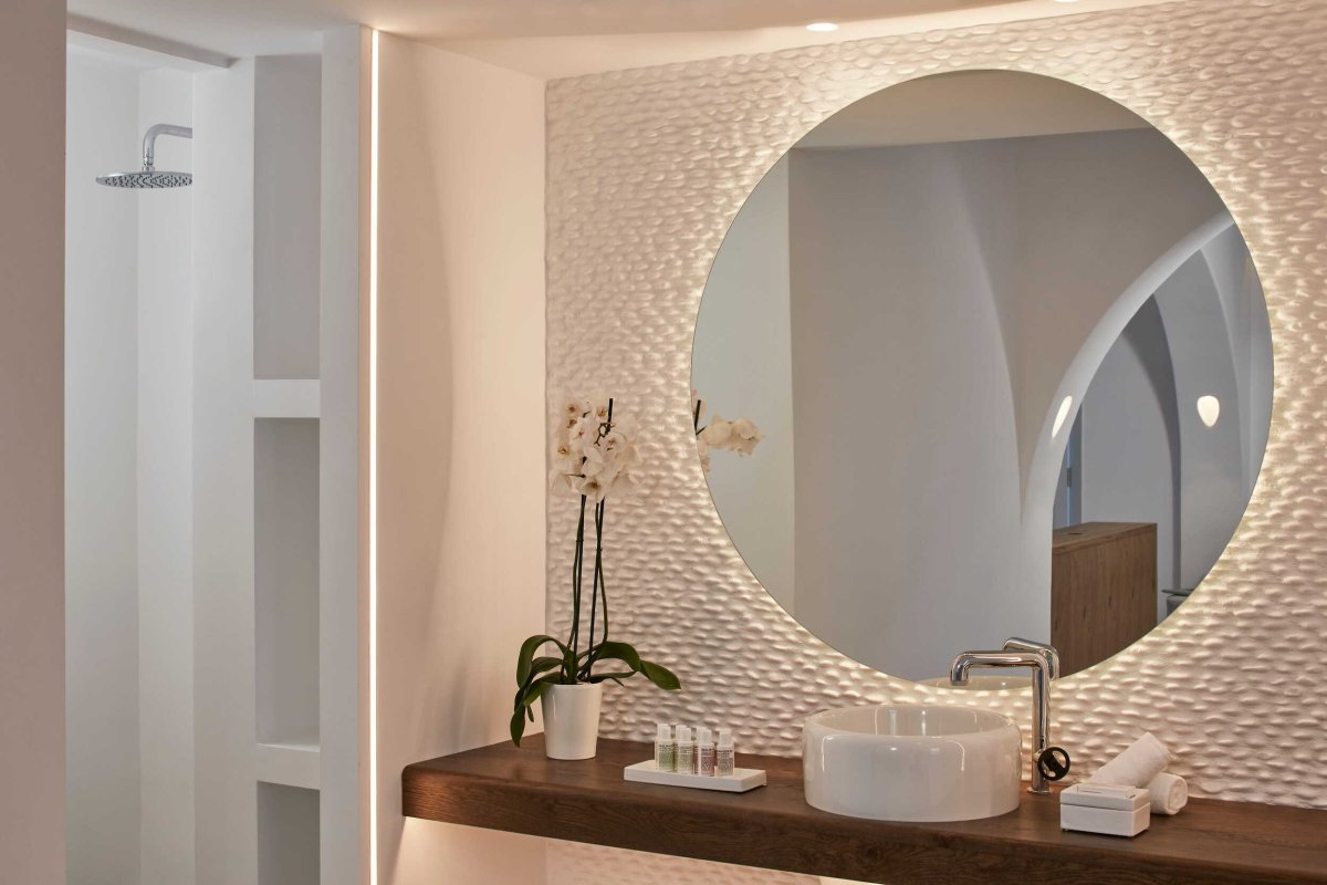 A modern, luxurious bathroom interior featuring a large round illuminated mirror, a textured white accent wall, a sleek wooden vanity with a white vessel sink, and an elegant potted orchid, reflecting the resort's high-end design