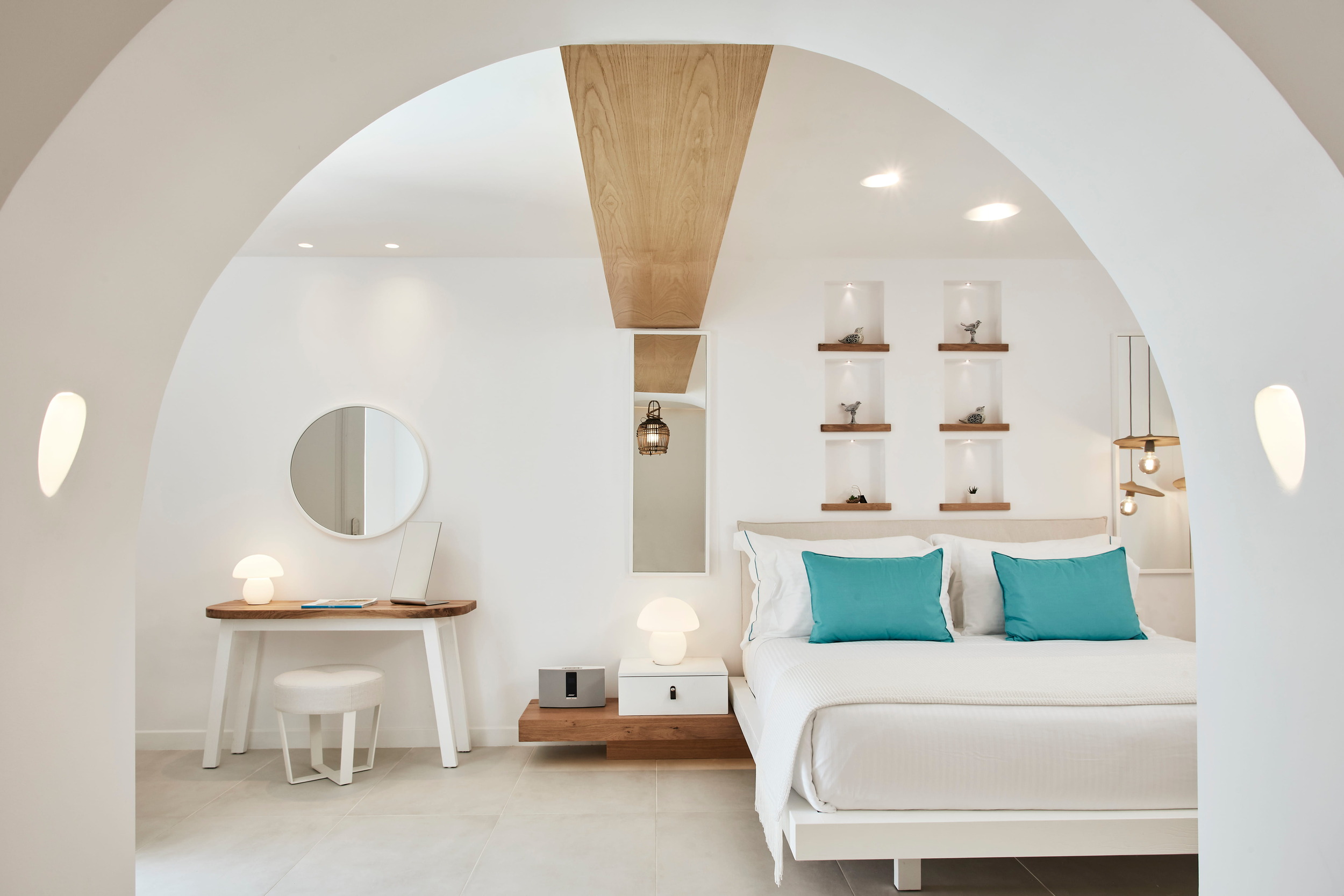 A view through a smooth white archway into a bright, modern luxury bedroom. The room features crisp white bedding with vibrant turquoise accent pillows, a sleek vanity desk with a round mirror, and warm wood architectural details including a ceiling beam and built-in display niches. This serene, stylish space beautifully represents the luxury accommodations awaiting guests at Nikki Beach Resort & Spa Santorini