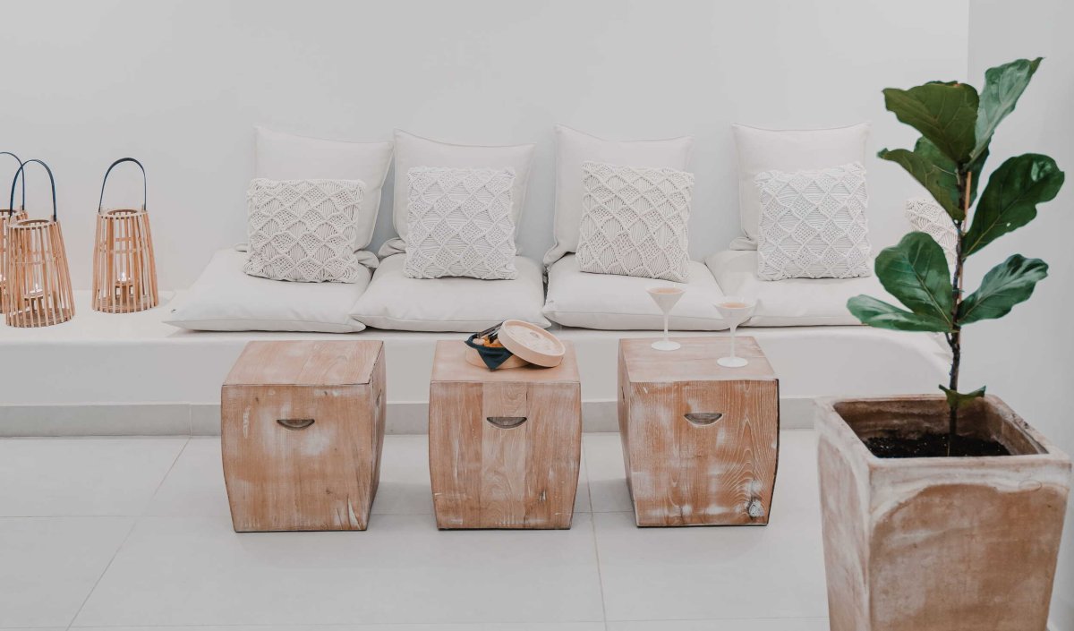 A chic, bohemian-inspired lounge area featuring white built-in seating with textured macrame pillows, three rustic wooden block tables holding cocktails, and elegant natural accents