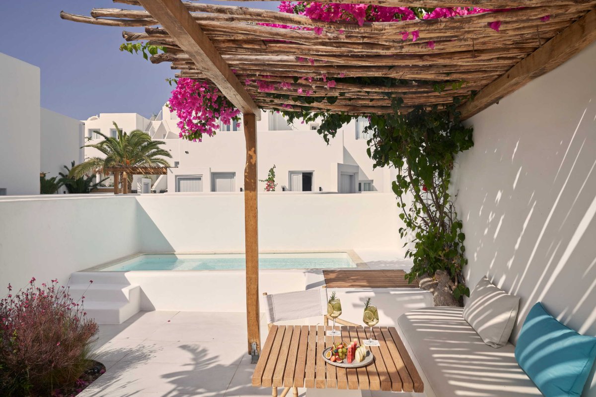 A luxurious private patio offering the perfect blend of relaxation, featuring a pristine plunge pool, a rustic wooden pergola draped in pink bougainvillea, and a cozy lounge area with drinks and a fruit platter