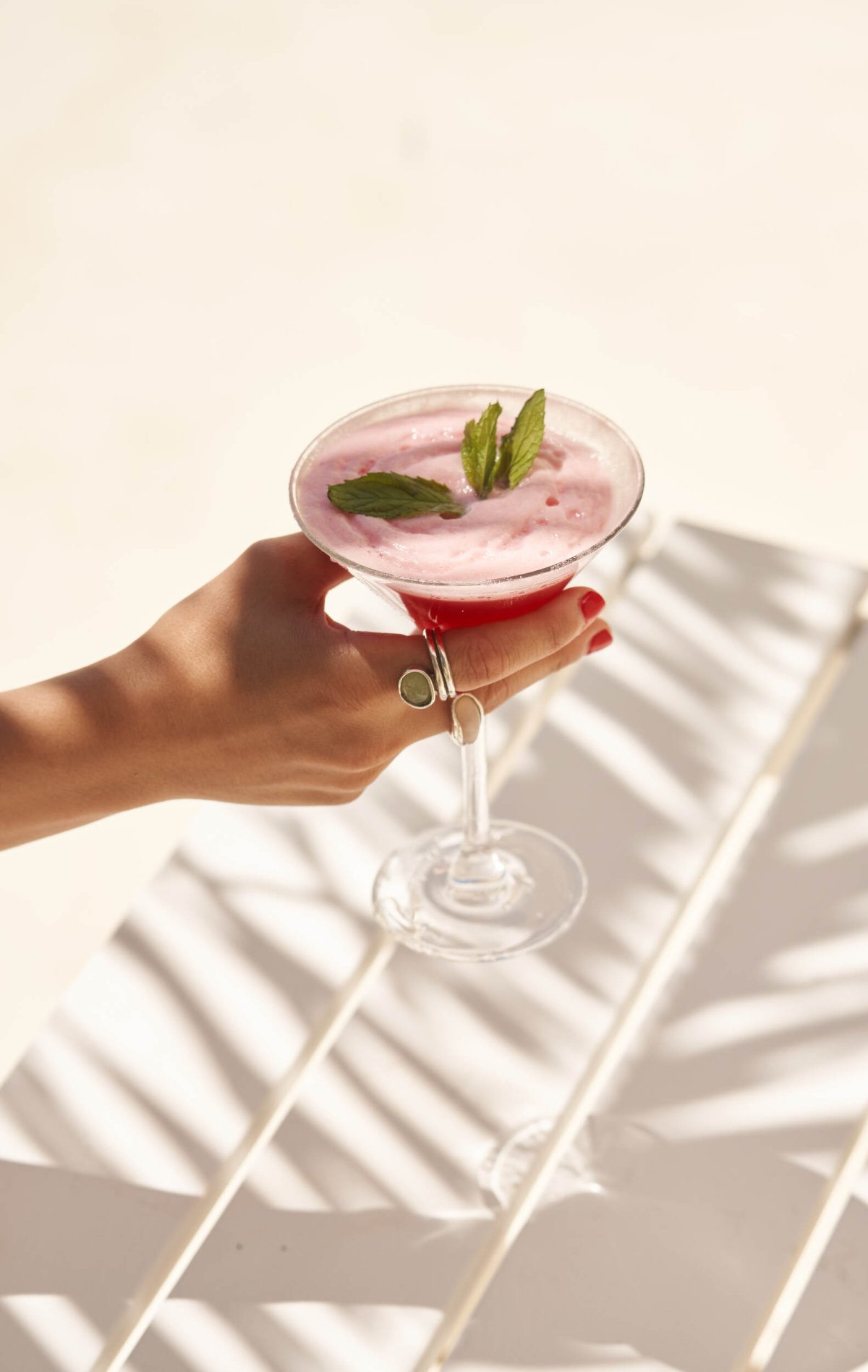 A hand holding a vibrant pink, frothy cocktail garnished with fresh mint over a white slatted table. Beautiful palm frond shadows add a warm, tropical touch to this refreshing drink