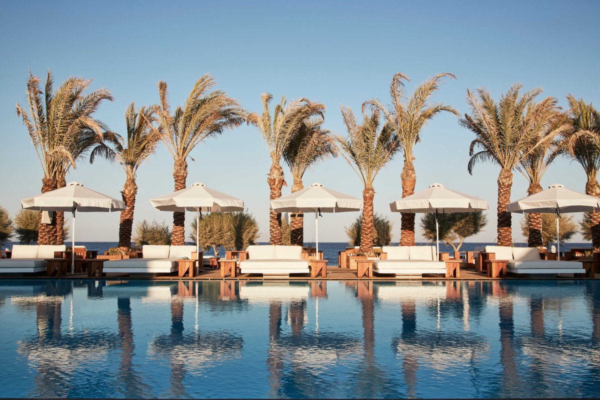 A stunning, expansive swimming pool perfectly reflecting a row of tall palm trees, plush white daybeds, and matching white umbrellas under a clear blue Santorini sky