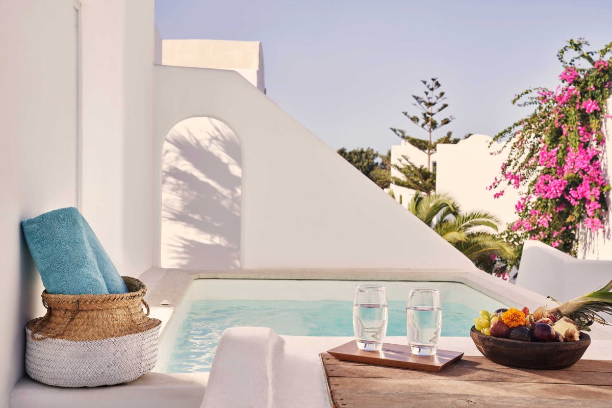A luxurious private outdoor jacuzzi framed by classic white architecture and bright pink bougainvillea, accompanied by fresh fruit, water, and a woven basket holding plush blue towels