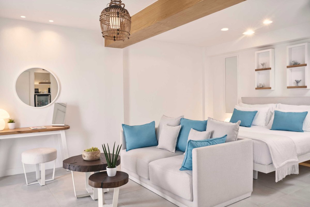A bright, modern hotel suite showcasing luxurious accommodations with crisp white bedding, a light grey sofa accented by turquoise pillows, natural wooden tables, and elegant built-in wall shelving