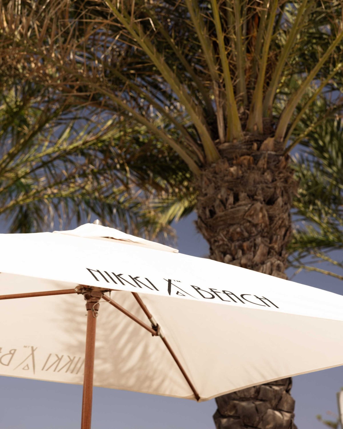 A signature white Nikki Beach umbrella positioned beneath the fronds of a lush green palm tree, capturing the tropical, barefoot luxury ambiance of the resort