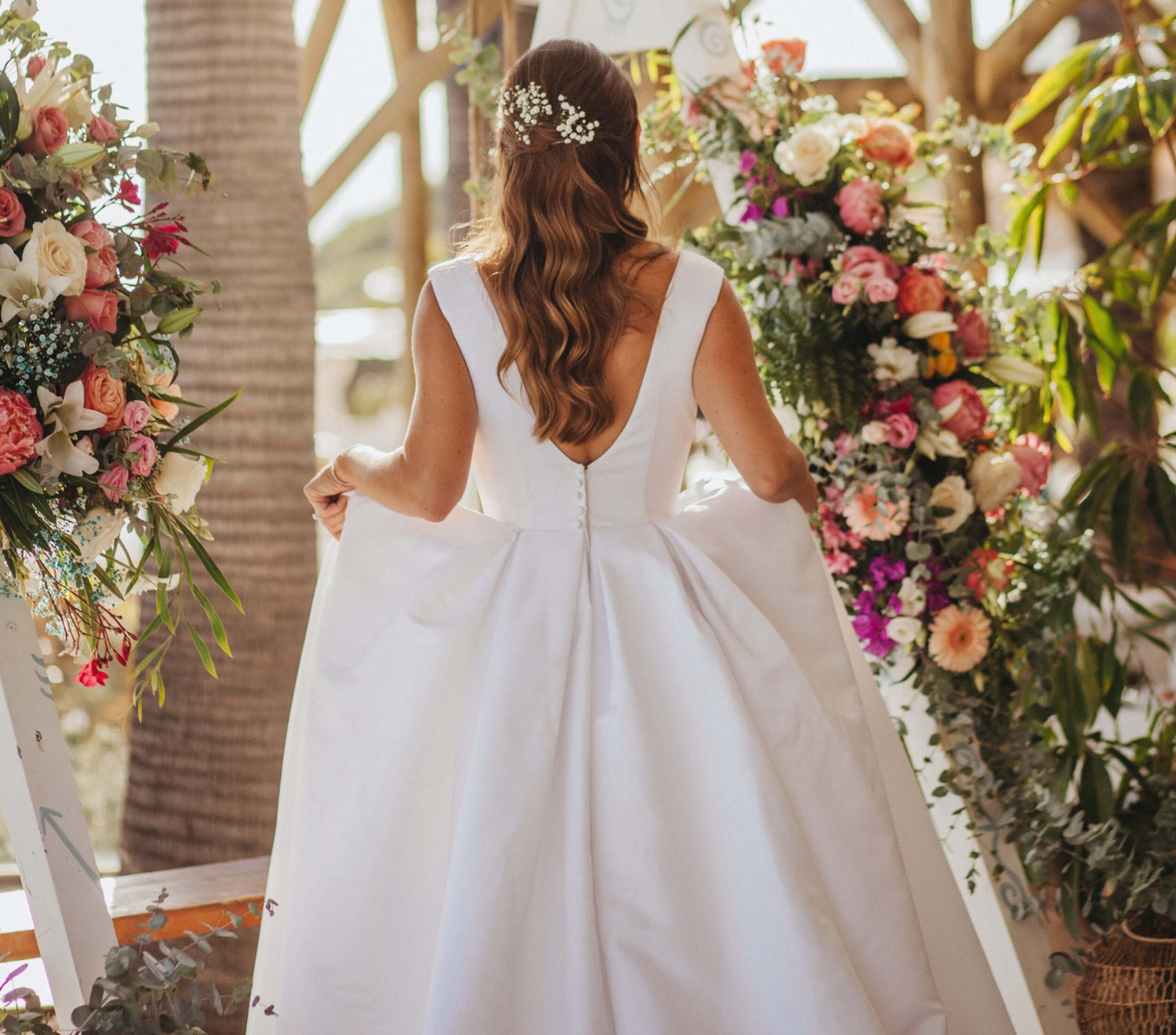 A candid shot of the bride walking away from the camera, showing her hair with a pearly hair clip. Holding her dress up slightly in front of her, she is walking between colourfully arranged flowers