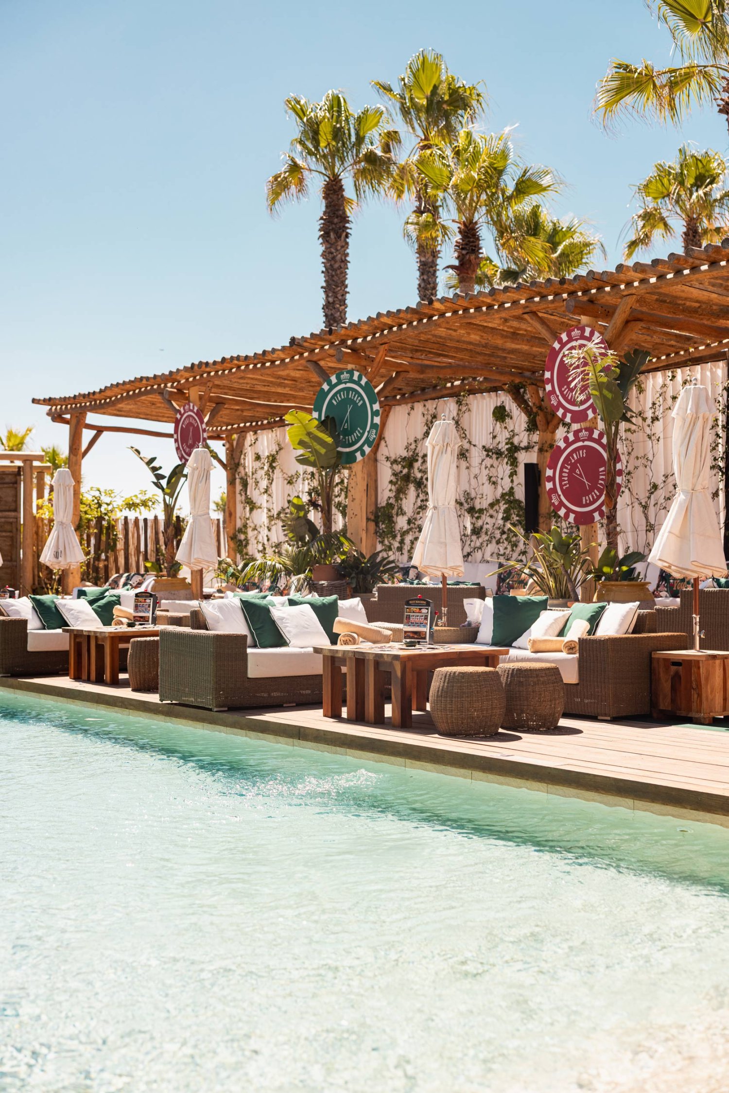 Outdoor pool area with sofas, stools and large coffee tables. A wooden canopy sits behind providing slight shade