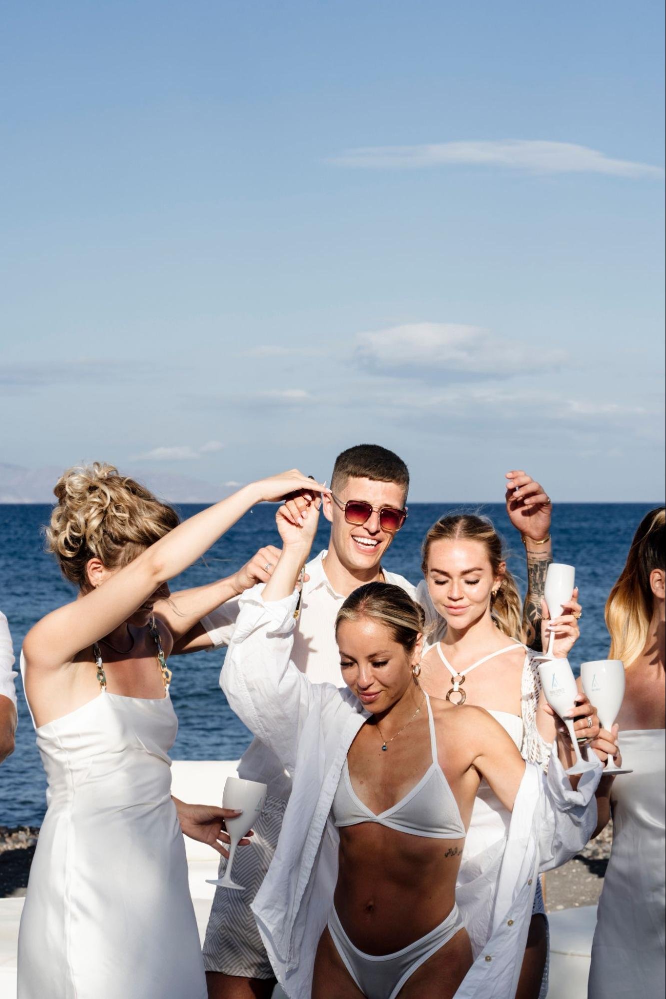 A group of guests dressed in stylish white resort wear dancing and celebrating together right next to the bright blue Aegean Sea. They are smiling, raising their arms, and holding signature white goblets, perfectly capturing the sun-soaked, high-energy daytime atmosphere of the Saturdance event