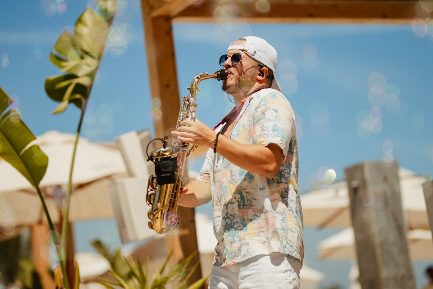 A live saxophonist playing energetically outdoors, surrounded by playful floating bubbles under a bright blue sky. He is wearing a tropical floral shirt, white pants, and sunglasses, perfectly capturing the pure energy and vibrant heartbeat of the weekly Saturdance event