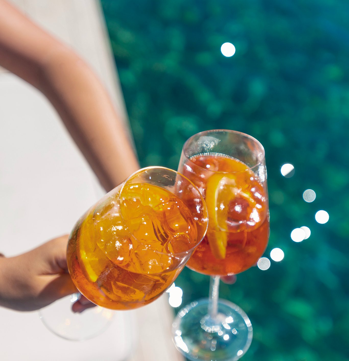 A close-up of two hands clinking large glasses filled with bright orange Aperol Spritz cocktails, garnished with fresh orange slices, over a sparkling blue swimming pool. The vibrant, refreshing drinks perfectly capture the relaxing midweek Spritz Escape experience