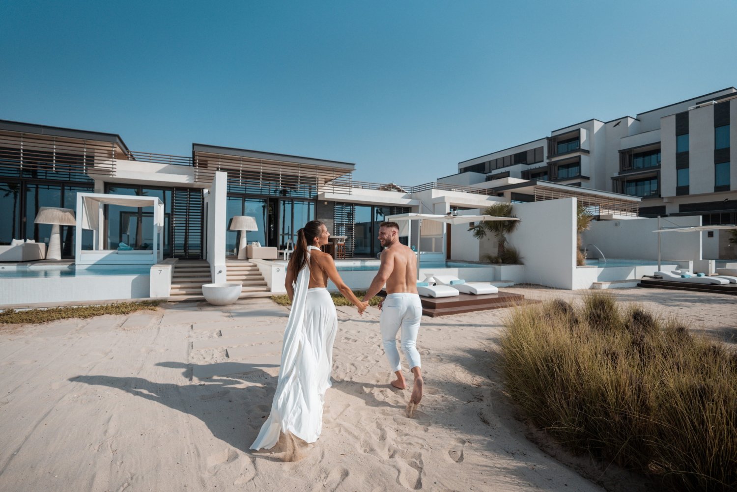 A couple enjoys a romantic walk on the beach, with a stunning resort behind them, evoking a sense of luxury and intimacy
