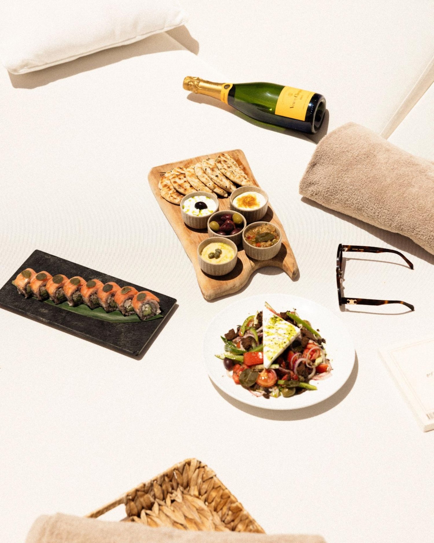 A top-down view of a Greek-inspired sharing menu laid out in the bright sun on a textured white daybed. The spread features a fresh Greek salad topped with a large block of feta, a wooden mezze board with pita bread and various dips, a plate of sushi rolls, and a bottle of chilled champagne resting next to a folded towel and sunglasses. This beautifully captures the relaxed, sun-drenched coastal charm and Mediterranean flavors of the Thursday Summer Club experience