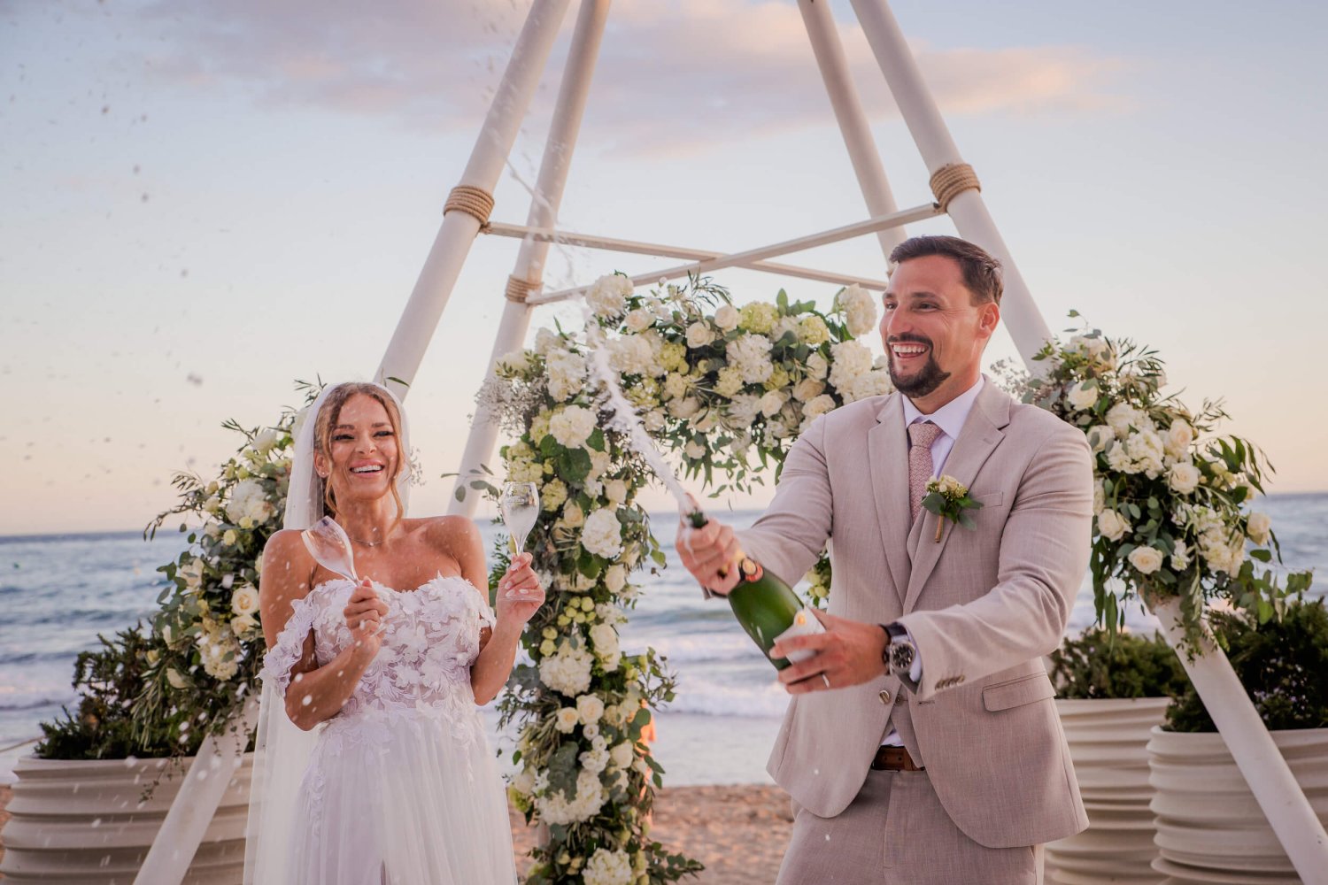 Newlyweds opening a champagne bottle after a beach wedding infront of the Nikki Beach teepee