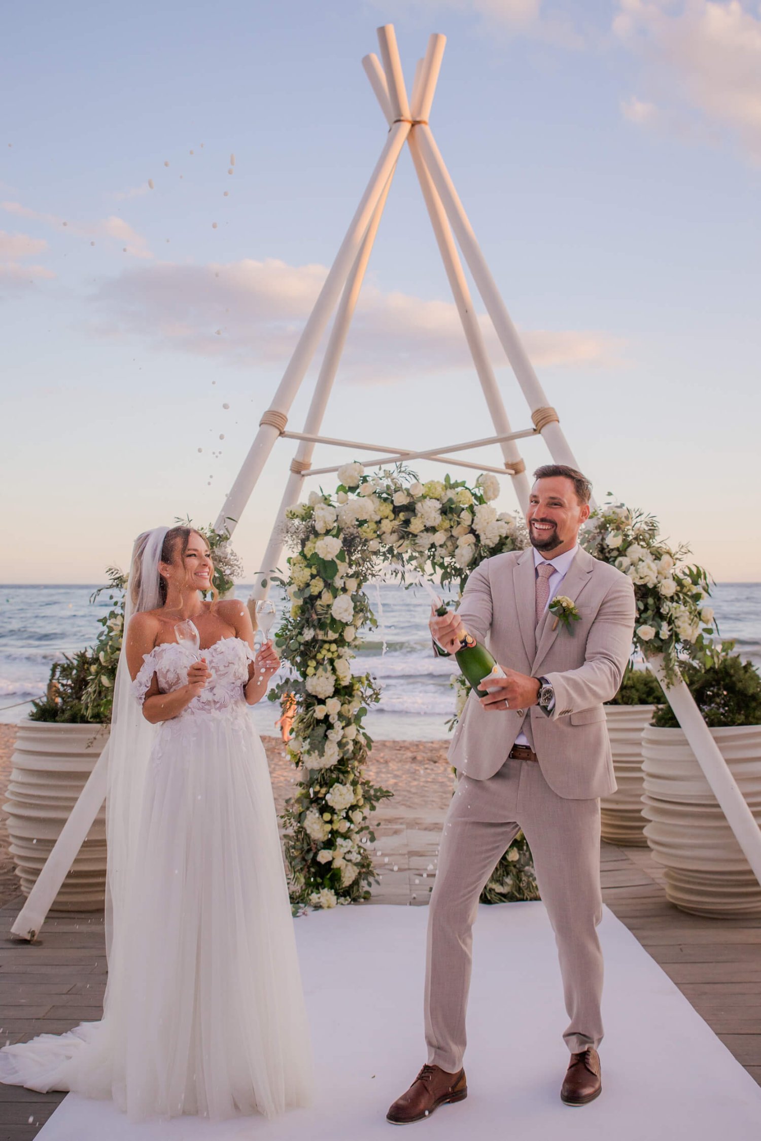 Newlyweds opening a champagne bottle after a beach wedding infront of the Nikki Beach teepee