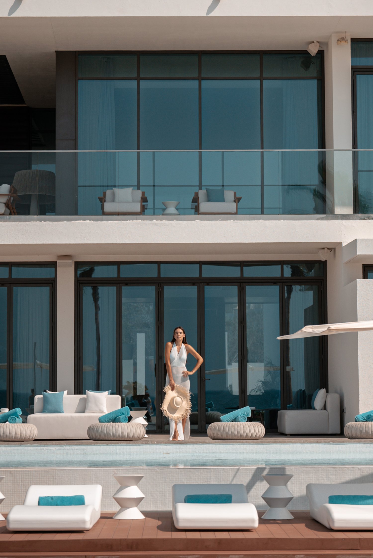 A women standing across the opposite side of the Ultimate Villas pool, holding a straw hat. Sun Loungers and cushions surround the edges of the pool
