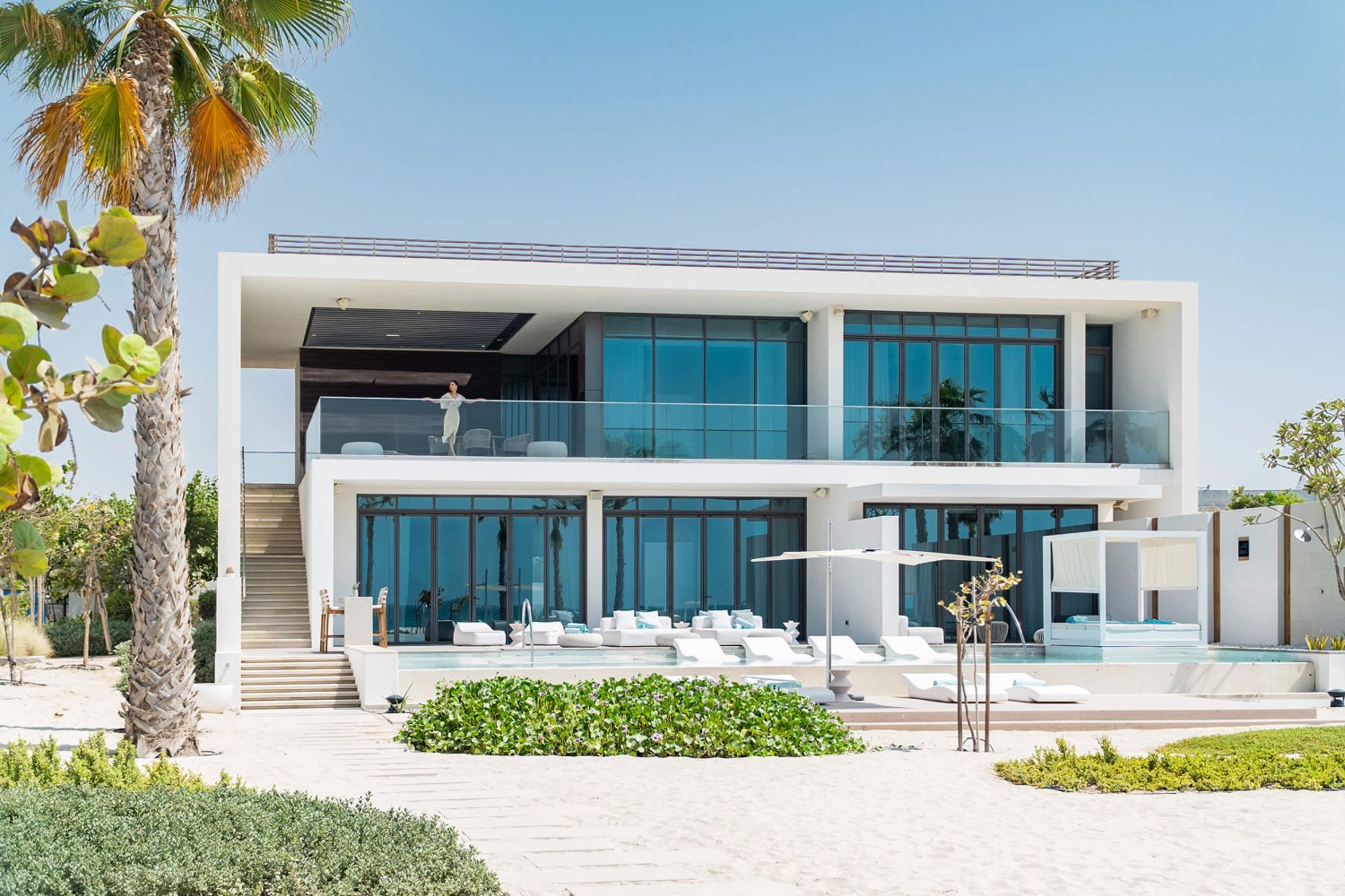A grand view of the ultimate villa at Nikki Beach Resort and Spa Dubai. A women stands on the second floor balcony overlooking the pool area and palm trees