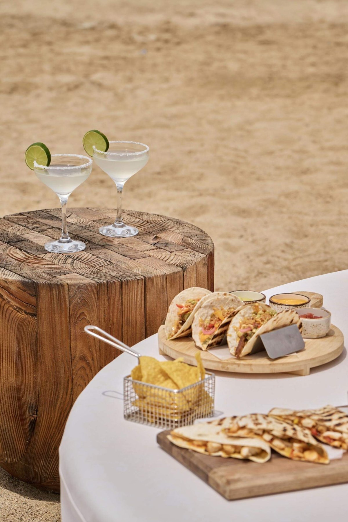 A table with a selection of tacos and nachos. Cocktails sit on a small wooden table beside the tacos