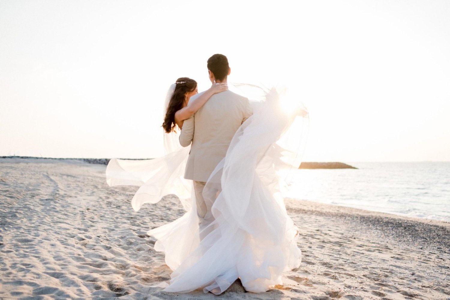 A romantic scene of a groom carrying his bride along a sandy shoreline bathed in golden sunlight. The bride's flowing white dress and long veil gracefully catch the ocean breeze as they walk near the water's edge. This beautiful moment perfectly captures the personal, beautifully composed beachfront wedding ceremonies offered at Nikki Beach Resort & Spa Koh Samui
